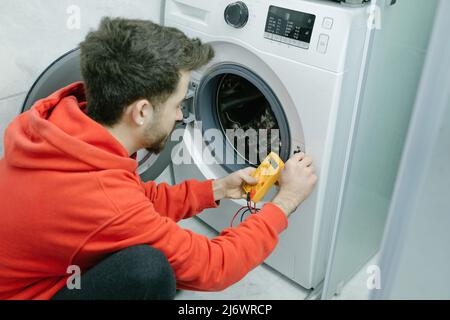Junger Mann mit rotem Hoodie, der zu Hause die Waschmaschine mit Digitalmultimeter überprüft. Stockfoto