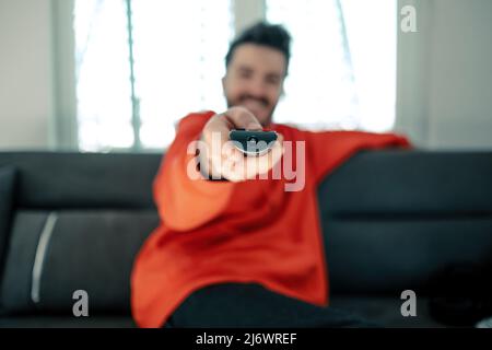 Junger Mann mit rotem Kapuzenpullover und Fernbedienung mit einem Lächeln auf dem Sofa. Fernsehen, während Sie zu Hause auf der Couch sitzen. Selektiver Fokus Stockfoto