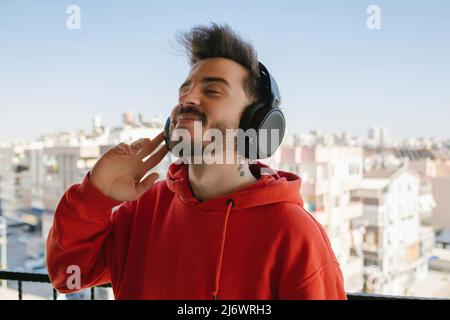 Fröhlicher junger Mann mit rotem Hoodie, der mit Kopfhörern Musik hört. Genießen Sie seine Lieblingsmusik. Kopfhörer hören und lächeln, während Sie auf dem Kopfhörer stehen Stockfoto
