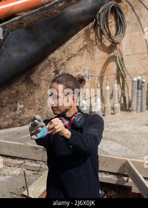 Junger Mann, der Schutz- und Sicherheitshandschuhe anziehen soll, umgeben von einer halben Konstruktion Stockfoto