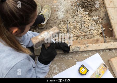 Draufsicht auf eine junge Frau, die auf einer von Beton umrundeten Baustelle einen Nagel in ein Holz geschlagen hat Stockfoto