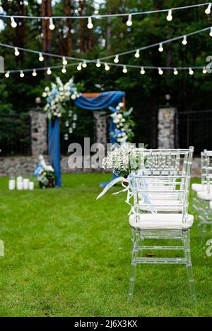 Ein Ort für eine Hochzeit im Freien, mit einem Sitzbereich für Gäste, dekoriert mit Stühlen. Rustikaler Holzbogen mit blauem Stoff, Blume dekoriert Stockfoto