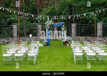 Ein Ort für eine Hochzeit im Freien, mit einem Sitzbereich für Gäste, dekoriert mit Stühlen. Rustikaler Holzbogen mit blauem Stoff, Blume dekoriert Stockfoto