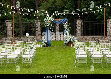 Ein Ort für eine Hochzeit im Freien, mit einem Sitzbereich für Gäste, dekoriert mit Stühlen. Rustikaler Holzbogen mit blauem Stoff, Blume dekoriert Stockfoto