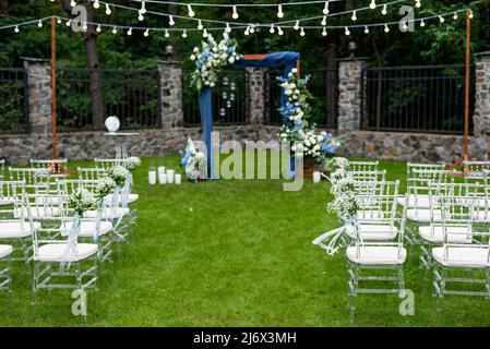 Ein Ort für eine Hochzeit im Freien, mit einem Sitzbereich für Gäste, dekoriert mit Stühlen. Rustikaler Holzbogen mit blauem Stoff, Blume dekoriert Stockfoto