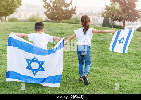 Kleine Kinder laufen mit israelischer Flagge. Rückansicht gesichtsloses kleines Mädchen und Junge, die mit der israelischen Flagge einen Hügel im Park hochlaufen. Stockfoto