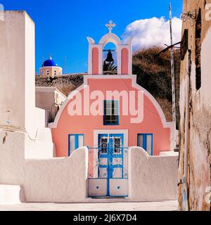 Oia, Santorini, Griechische Inseln, Griechenland - hübsche rosafarbene griechisch-orthodoxe Kirche mit Glockenturm und blauer Kuppel hinter der Kirche in strahlend heller Sonnensegel Stockfoto