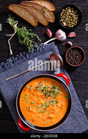 Dicke Süsskartoffelsuppe mit Gemüse in rotem Topf auf dunklem Holztisch mit Kräutern, Pfefferkörnern, Kürbiskernen und Roggenbrotscheiben, vertikal Vie Stockfoto