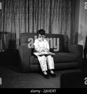 1950s, ein historischer Junge mit Pyjamas und Pantoffeln, der auf einem kleinen Sofa sitzt und vor dem Schlafengehen ein Bilderbuch ansieht, England, Großbritannien. Stockfoto