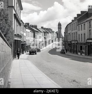1950s, historisch, Enniskillen, Nordirland, Großbritannien, Es zeigt eine nahe verlassene Hauptstraße mit nur ein paar Einheimischen, die auf dem Bürgersteig spazieren. Ein Gabelstapler mit Milchurnen wird hochgeparkt. Am Ende der Straße ist der Uhrenturm des Rathauses aus dem Jahr 1901 zu sehen. Zu den Namen in den Geschäften gehören eine Bar, William Blake, und ein Metzger und Fleischlieferant Peter Burns. Das Nummernschild auf einem der Autos, die in der verlassenen Straße, IL 2202, geparkt sind, während auf der anderen Straßenseite ein anderes Auto aus einer noch früheren Zeit geparkt ist und dieses das Nummernschild HMT 275 trägt. Stockfoto
