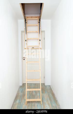 Klappbare Holztreppe im Dachgeschoss auf leerem weißen Hausflur hochziehen. Enger Flur mit weißen Wänden und blauem Laminatboden. Stockfoto