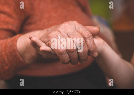 Die Hände des Enkels halten die faltige Hand einer kranken älteren Großmutter zu Hause. Das Konzept von Liebe und Fürsorge. Langsame Bewegung. Stockfoto