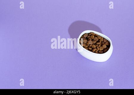 Draufsicht auf weiße Schale mit trockenem Kibble Katzenfutter auf lila, lila Hintergrund Stockfoto