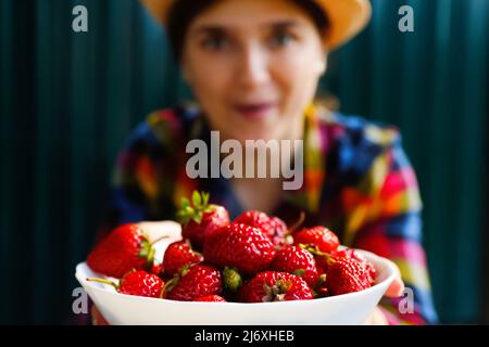 Unschärfe junge Frau in einem Hut und Hemd lächelt und zeigt eine weiße Schüssel mit Erdbeeren. Dunkelgrüner Stahlhintergrund. Sommeressen, Obst. Sommerzeit. Z. B. Stockfoto