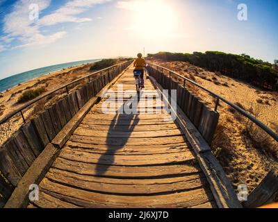 Eine Frau fährt mit dem Fahrrad auf einem Holzweg durch den Sand am Strand von Isla Cristina Stockfoto