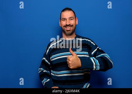 Bärtiger lateiner, gekleidet in gestreiften Pullover, zeigt Daumen nach oben, lächelt und schaut auf die Kamera, isoliert auf blauem Hintergrund. Stockfoto