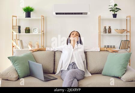 Entspannte Frau atmet frische Luft ein, während sie auf dem Sofa im Wohnzimmer mit Klimaanlage sitzt Stockfoto