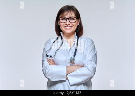 Porträt einer lächelnden Ärztin, die eine Brille in weißer Uniform trägt, mit Stethoskop, das die Kamera anschaut, und einer selbstbewussten Doc mit gekreuzten Armen im Lichtstudio Stockfoto
