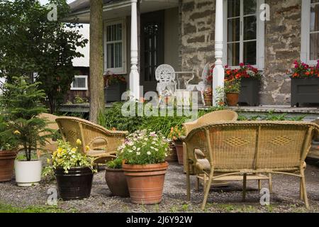 Topfpflanzen und Korbmöbel in landschaftlich gestaltetem Vorgarten vor dem 1700s Canadian Stil Feldsteinhaus im Sommer. Stockfoto