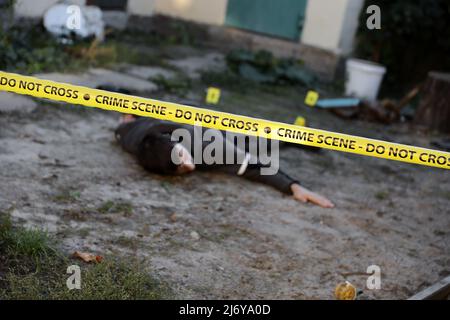 Opfer eines gewalttätigen Verbrechens in einem Hinterhof des Wohnhauses am Abend. Leiche eines toten Mannes unter dem gelben Polizeistreifen-Klebeband und Beweismarkierungen für Verbrechen Stockfoto