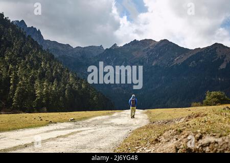 Rückansicht des asiatischen Wanderreisenden Rucksacktouristen, der auf einer unbefestigten Straße in Richtung Berge und Wald geht Stockfoto