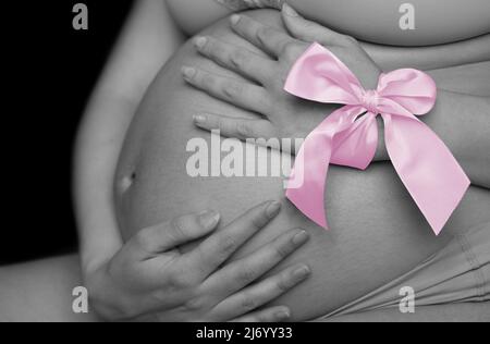 Bauch einer Schwangeren von der Seite in schwarz und weiß. Die Frau hält einen rosa Bogen auf der Hand Stockfoto