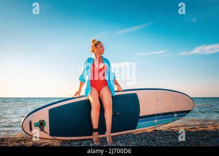 Mollige schöne kaukasische Frau posiert mit einem Schmollenbrett. Im Hintergrund ist der Himmel das Meer. Das Konzept der sportlichen Erholung. Stockfoto