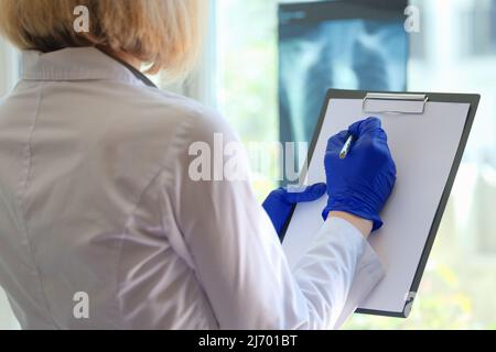 Die Hand des Arztes schreibt Notizen auf das Clipboard in der Medizin und schaut durch den Röntgenfilm Stockfoto