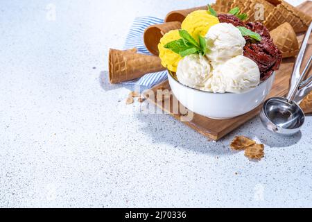 Zitronen-, Schokolade- und Vanilleeis. Große Schüssel mit Eisschaufeln, auf weißem Küchentisch-Hintergrund mit Waffelkegeln, Kopierfläche Stockfoto