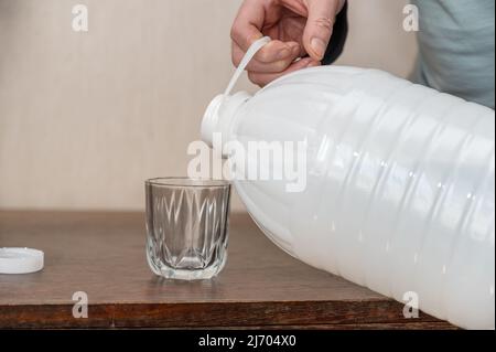 Ein Mann gießt Trinkwasser in ein Glas. Große geöffnete weiße Plastikflasche mit frischem Wasser. Stockfoto