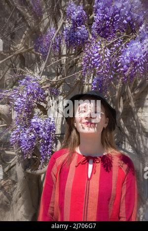 Die hübsche junge Frau steht neben der Glyzinie. Mädchen genießen Frühling Zeit. Outfit für den Frühling. Stockfoto