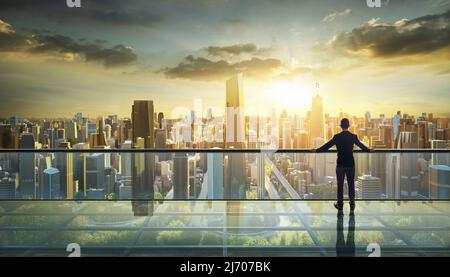 Geschäftsmann, der auf einem offenen Dachbalkon steht und den Blick auf den Sonnenaufgang in der Stadt beobachtet. Geschäftsambitionen und Vision-Konzept Stockfoto
