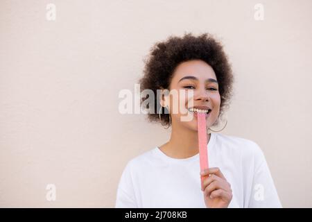 Lächelnde afroamerikanische junge Frau, die süßes Gelee isst, Luftblasenband auf Weiß Stockfoto