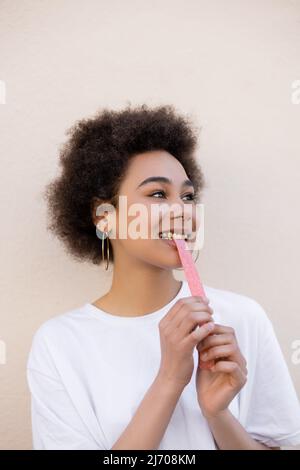 Erfreut afroamerikanische junge Frau, die süßes Gelee-Luftblasenband auf Weiß isst Stockfoto