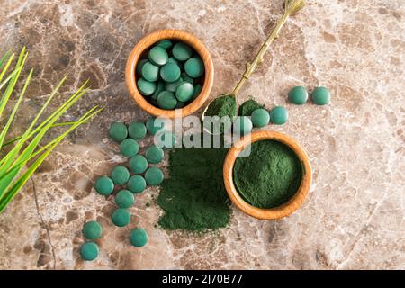 Grünes Spirulina-Pulver und Tabletten in Holzschalen auf braunem Marmorboden. Ein beliebtes Superfood für Vegetarier und Sportler. Draufsicht Stockfoto