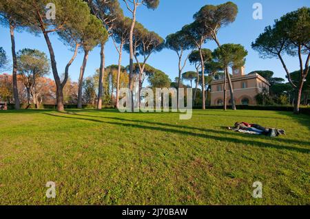 Ein junges Paar liegt auf dem Rasen im Park der Villa Borghese, im Hintergrund das Uhrgebäude (Casino dell'Orologio), Rom, Latium, Italien Stockfoto