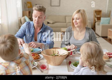Glückliche kaukasische Familie mit Kindern, die am Esstisch im Wohnzimmer sitzen und leckeres Mittagessen essen, Vater gibt Sohn Stück hausgemachten Kuchen Stockfoto