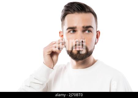 Ein hübscher Mann mit Bart in weißer Kleidung, der schnurlose Ohrstöpsel auf weißem Hintergrund verwendet Stockfoto