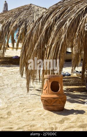 Lehmurne am Strand, vor dem Hintergrund von Sonnenschirmen, die an sonnigen Tagen mit trockenen Palmenblättern bedeckt sind. Vertikales Foto. Nahaufnahme. Selektiver Fokus. Stockfoto