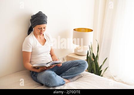 Eine Frau mittleren Alters sitzt auf einem Bett in ihrem Zimmer und liest ein Buch. Stockfoto