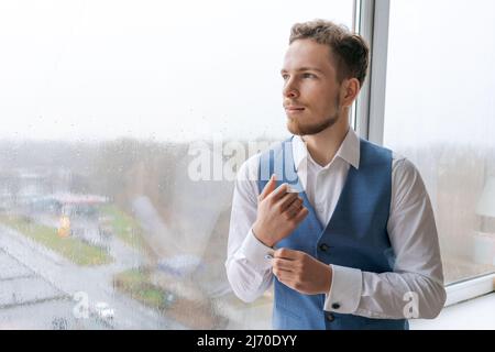 Portrait ein arroganter, scharf gekleideter Mann in blauem Anzug korrigiert Knopf auf Manschettenknöpfen sein Hemd, bereitet sich auf das Date Meeting vor, schaut lächelnd zum Fenster hinaus und erwartet eine erfolgreiche Transaktion Stockfoto