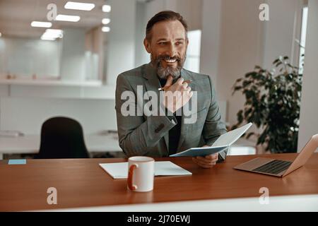 Ein glücklicher bärtiger Mann hielt ein Notizbuch, während er an seinem Schreibtisch saß Stockfoto