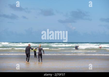 Eine Mutter und ein Vater halten die Hände mit ihrem Kleinkind, das im Meer bei Mawgan Porth in Cornwall paddelt; ein Urlaub für Familien in Cornwall. Stockfoto