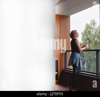 Quadratische Aufnahme einer kaukasischen Frau, die auf ihrem Balkon steht und eine Tasse Tee oder Kaffee in der Hand hält. Die Hälfte des Fotos ist weiß. Speicherplatz kopieren. Hochwertige Fotos Stockfoto