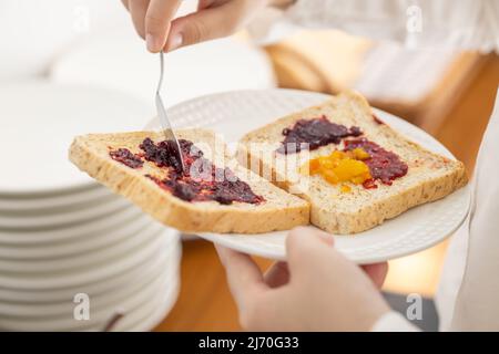 Tragen Sie süße Beerenobst-Marmelade auf ein in Scheiben geschnittenes Vollkornbrot zum Frühstück mit einer energiereichen Mahlzeit am Morgen auf Stockfoto