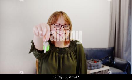 Junge rothaarige Frau zeigt die Schlüssel zu ihrer neuen Wohnung und schaut lächelnd auf die Kamera. Hochwertige Fotos Stockfoto