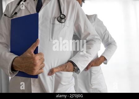 Selbstbewusste medizinische Fachkräfte, die mit den Händen in den Taschen stehen, tragen in der Mitte der Ärzte Laborkittel, sie sind gegen Weiß Stockfoto