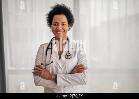Porträt einer selbstbewussten lächelnden Ärztin, Doktor steht mit gekreuzten Armen am Fenster, sie trägt Labormantel und Stethos Stockfoto