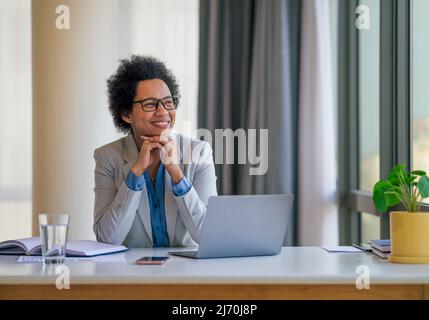 Eine nachdenkliche lächelnde Geschäftsfrau, die während der Arbeit am Laptop wegschaut, eine Unternehmerin trägt formelle Kleidung am Schreibtisch, sie denkt im Büro nach. Stockfoto