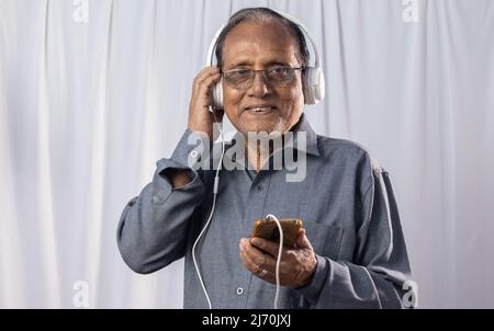 Eine indische alte Person, die Musik mit Smartphone und Kopfhörer auf weißem Hintergrund hört Stockfoto